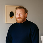 Bearded man with red hair wearing a dark blue sweater standing in front of a beige wall with abstract art.